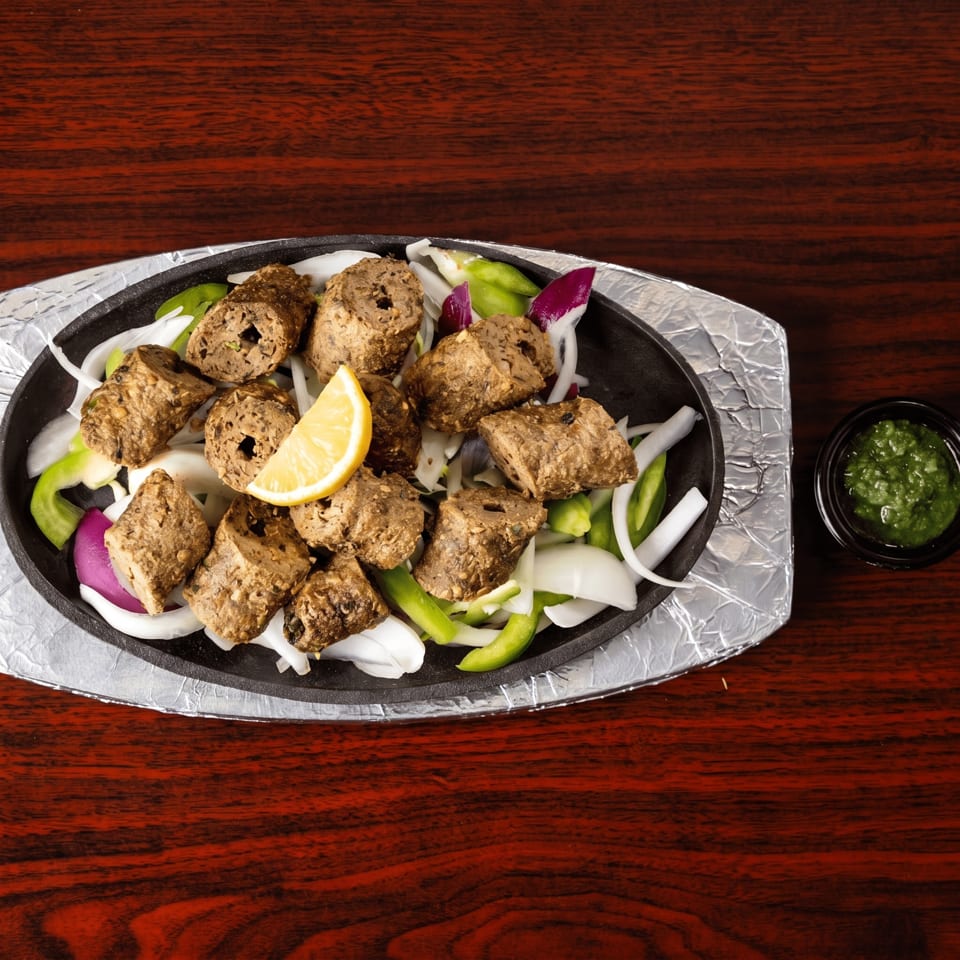 Sizzling platter of seekh kebabs with sliced onions, green peppers, and a lemon wedge.