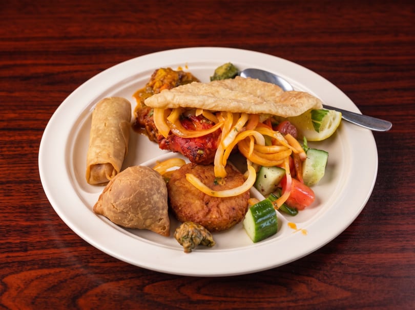 A white plate filled with various Indian dishes, including samosas, pakoras, and a flatbread.