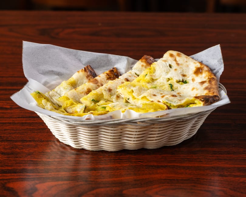 A white basket filled with pieces of golden-brown naan bread, some with yellow filling, on a wooden table.