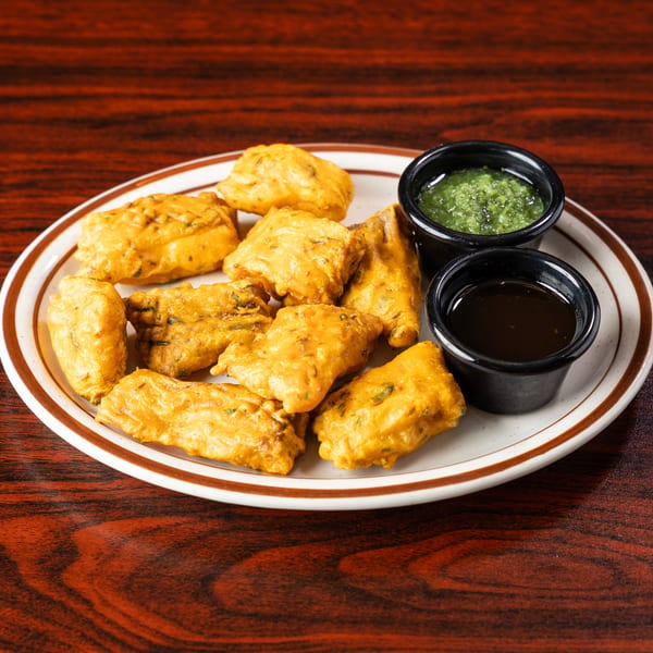 A plate of golden-brown fried pakoras with two small bowls of green and dark dipping sauces.