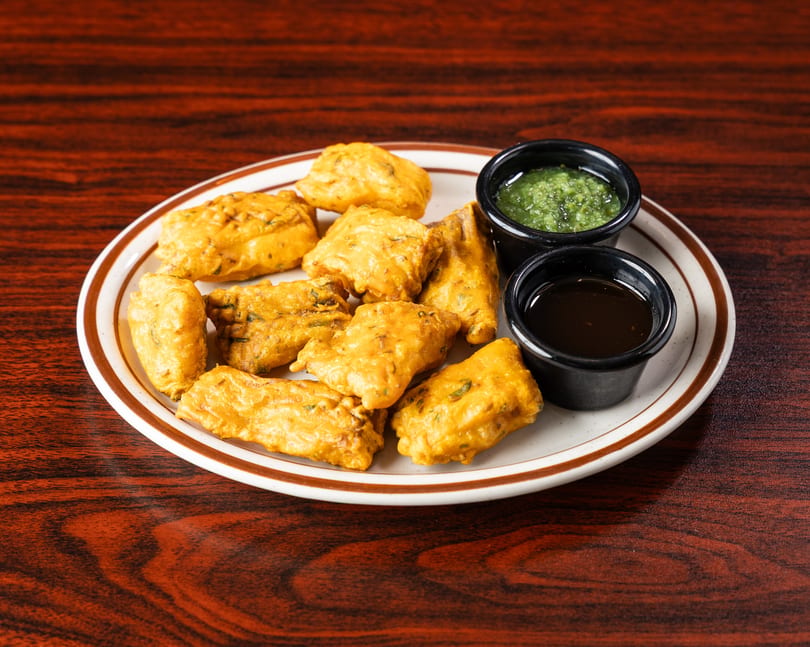 A plate of golden-brown fried pakoras with two small bowls of green and dark dipping sauces.