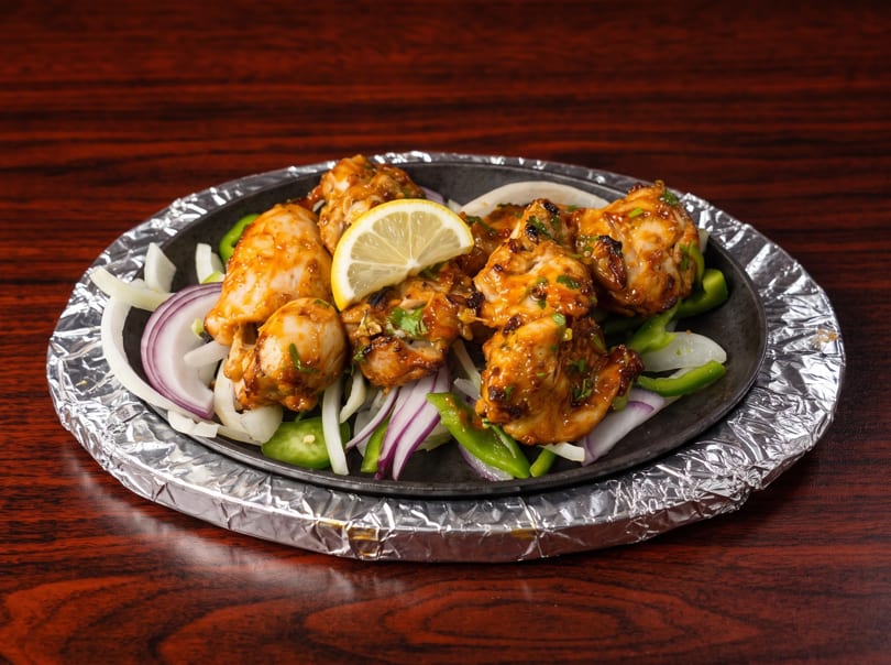 Sizzling chicken tikka with onions, green peppers, and a lemon slice on a foil-wrapped platter.