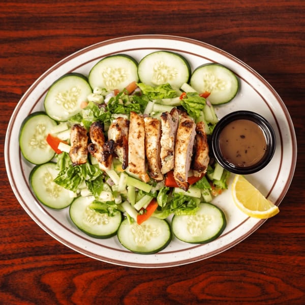 Grilled chicken salad with cucumbers, lettuce, red peppers, and a side of dressing and lemon.
