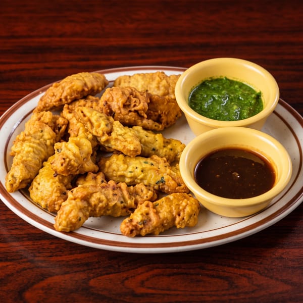 Golden brown fried pakoras on a white plate with two dipping sauces on a wooden table.