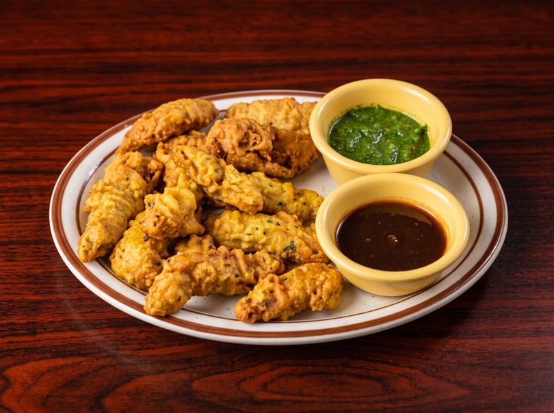 Golden brown fried pakoras on a white plate with two dipping sauces on a wooden table.
