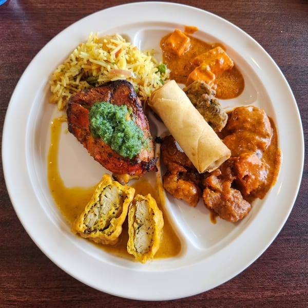 A white partitioned plate holds various Indian dishes, including rice, curries, chicken, and a spring roll.